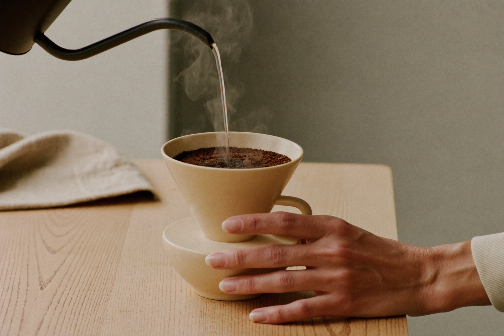 Hænder hælder varmt vand over malet kaffe i en keramisk dripper på en lys egetræsdisk